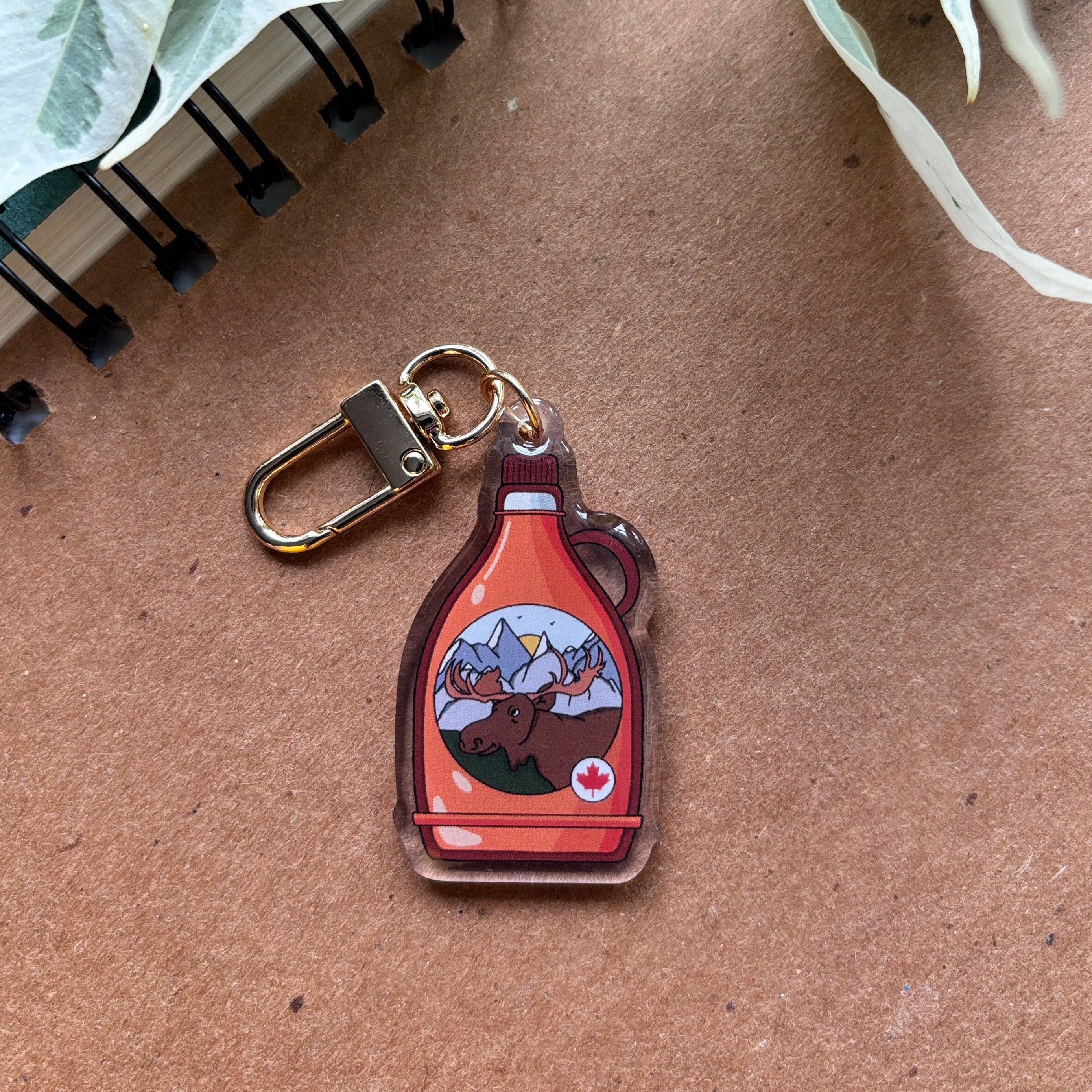 Keychain with a design of a maple syrup bottle on a brown surface with green leaves.