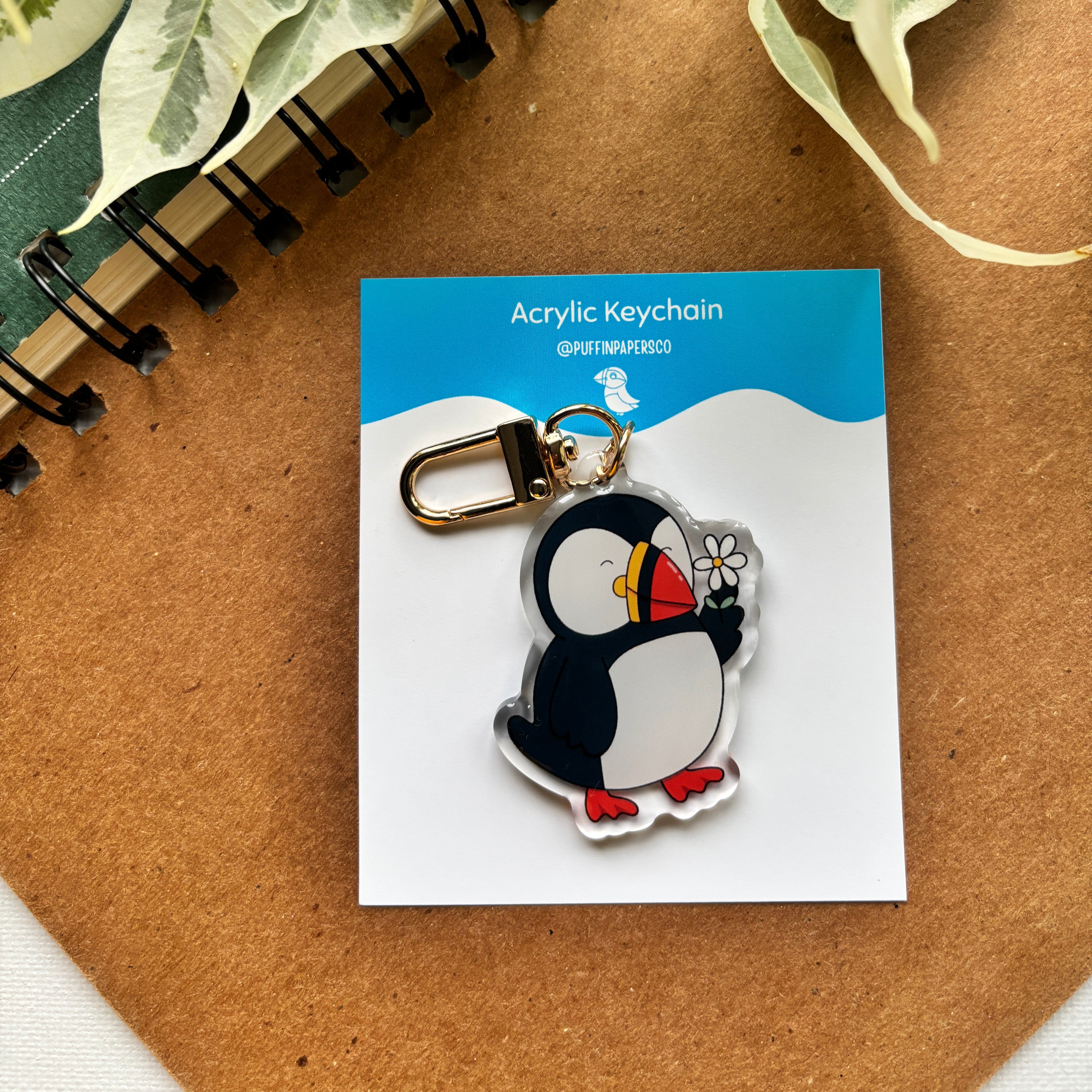 Penguin acrylic keychain on a card with a notebook and leaves in the background